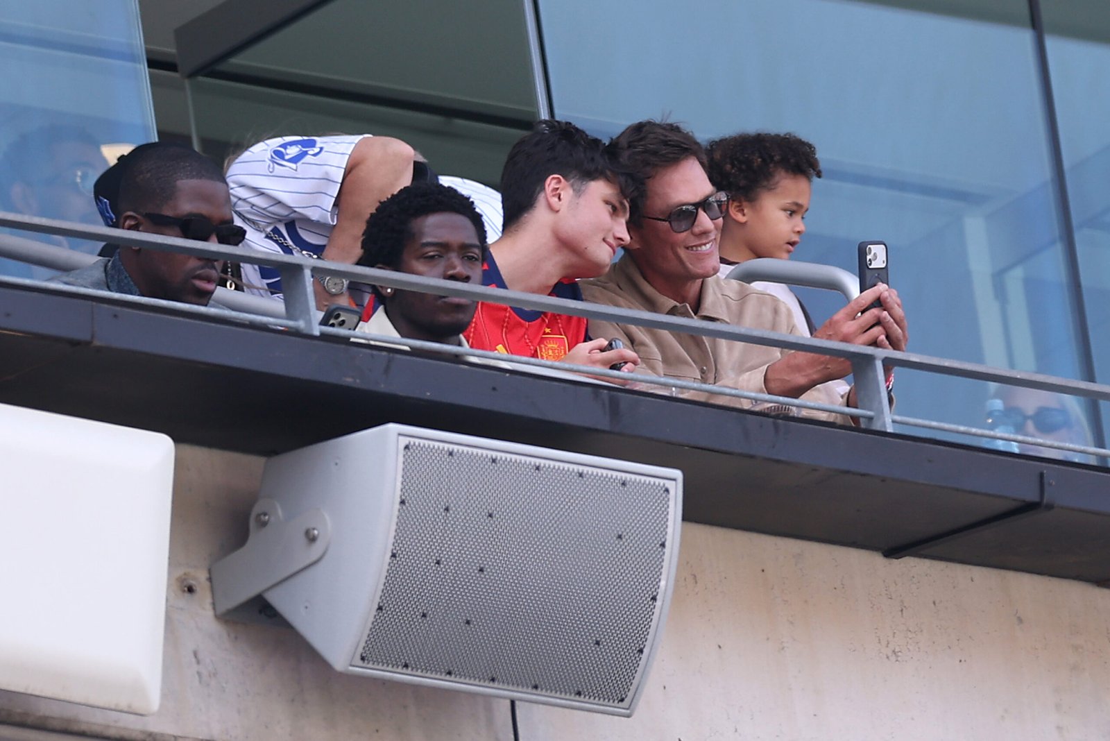 Tom Brady smiles and captures a selfie with his young son peering over his shoulder from their stadium balcony suite, enjoying football's biggest night at the FIFA World Cup 2026 Final match.