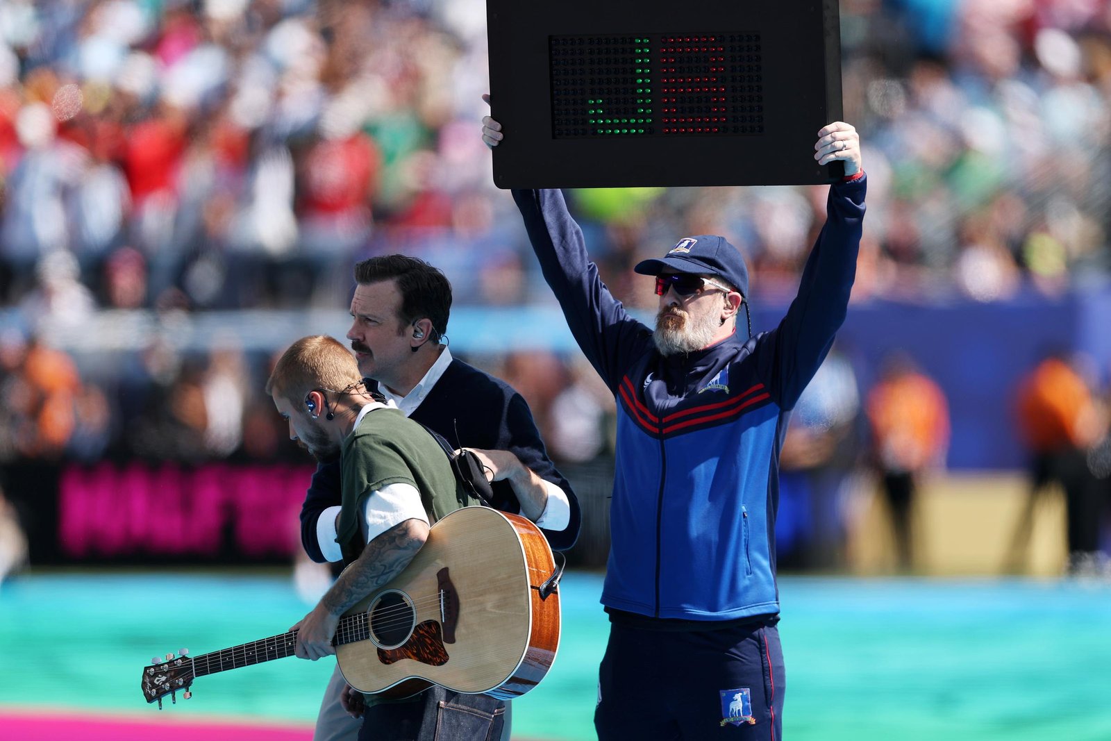 A stadium crew member holds an electronic board displaying a giant "J" and "US" during Justin Bieber's halftime appearance, adding a branded visual to the musician's low-energy acoustic interlude.