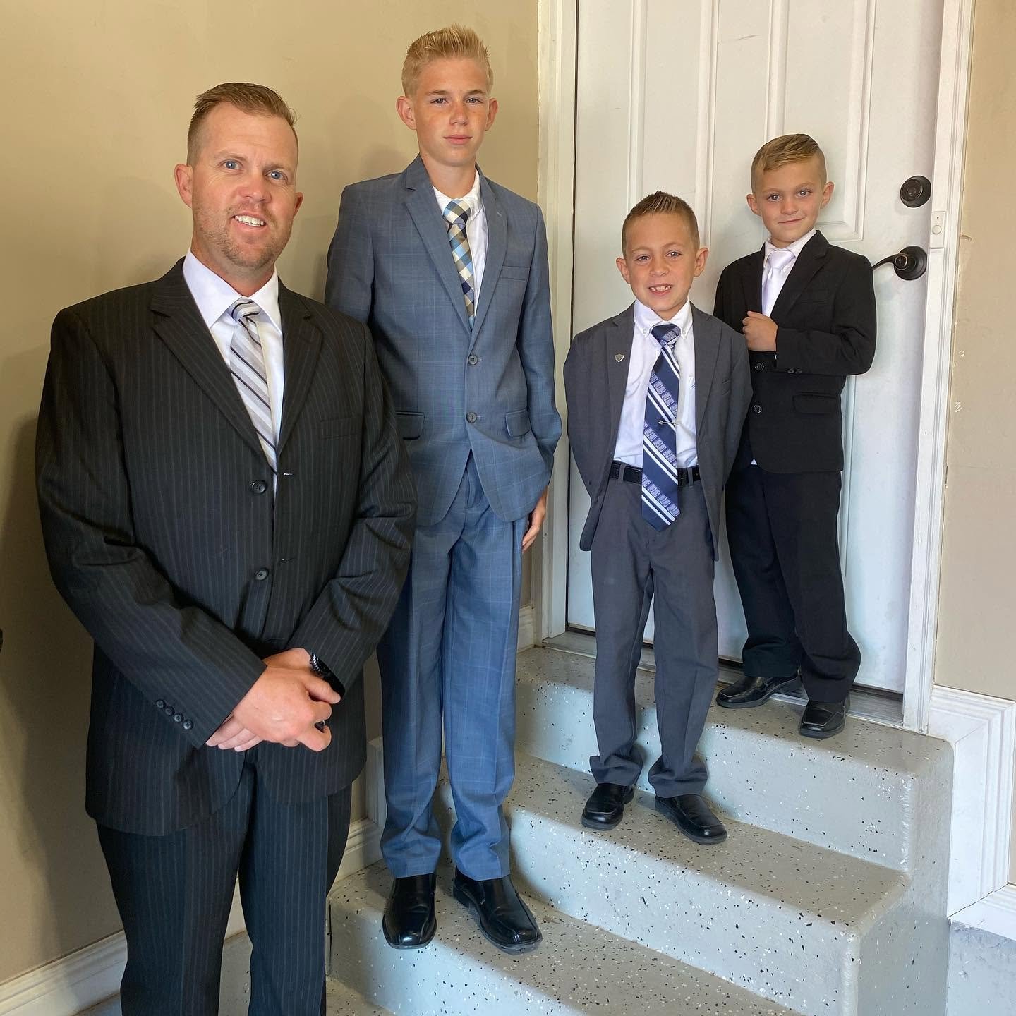 Spencer stands at the forefront with hands respectfully clasped, posing indoors with his three handsome sons, Reid, Thayne, and Gage, on a nearby stairway. The quartet looks unified and sharp in their tailored suits, representing a legacy of strength and unity.