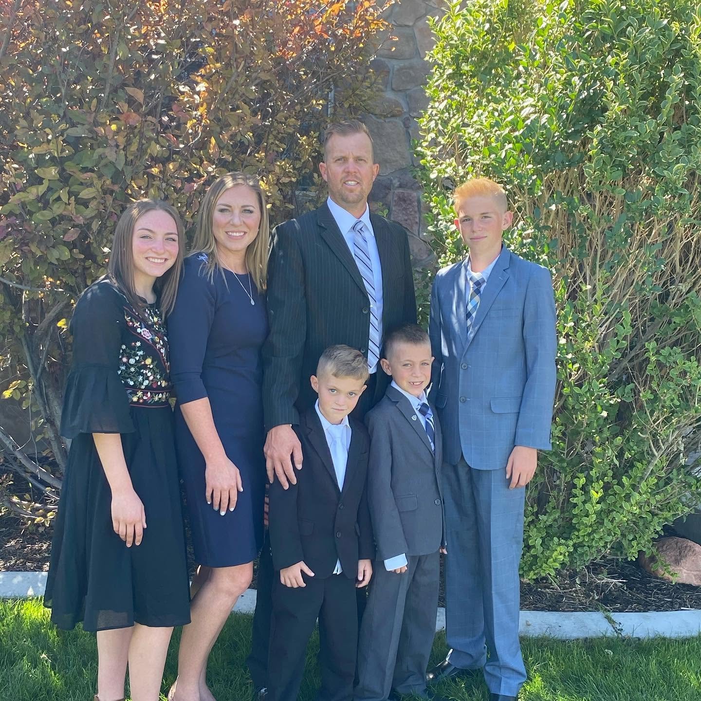 The entire Long family poses formally outdoors against a backdrop of green foliage and a stone wall. Katrina stands elegant in navy, Lydia in a detailed black dress, and Captain Spencer looks sharp alongside his three sons, Reid, Thayne, and Gage, all dressed in their sharpest suits.
